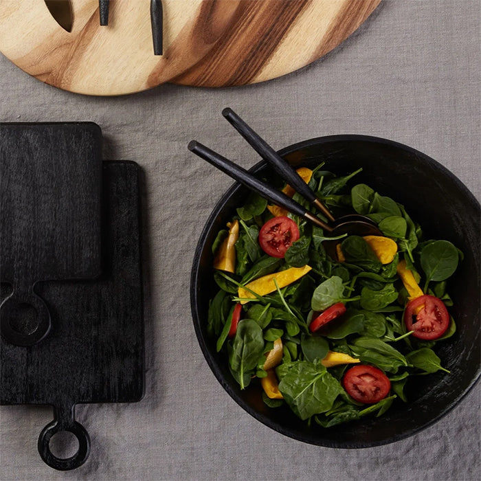 Black mango wood serving bowl filled with fresh salad, placed on a neutral tablecloth with black cutting boards.